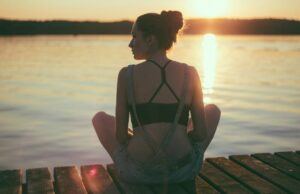 a lady in black shorts and black sports bra sitting cross legged with her back towards the camera looking out over a sunset and lake