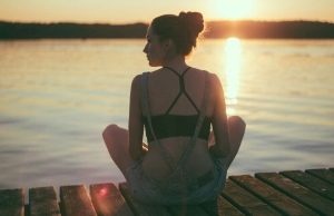 a lady in jean shorts and black sprots bra relaxing on a wooden pier in front of a lake watching a sunset