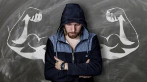 a light skinned man in a blue jacket with his hood up looking down while standing in front of a background that has muscly arms outstretched